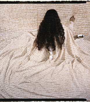 A figure with long, dark, curly hair is seated facing away from the viewer, draped in material covered in calligraphy. The same material covers the background of the image, and the figure's right hand is raised, holding a pen, writing on the material.
