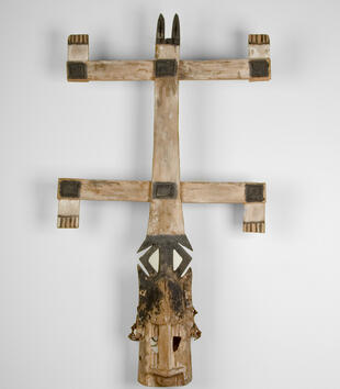 The face of a wooden mask with a long nose and pointed ears is topped with a wooden, vertical structure with upper and lower cross bars. There are notches at the ends of the crossbar and a pair of black ears carved atop the vertical pole.