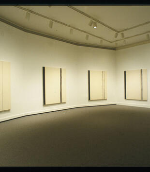A series of large white and black paintings hangs on the walls of a white gallery space. The white canvases are painted with black lines of differing thicknesses. 