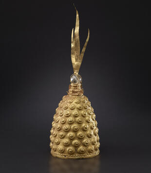 A tall, gold oblong crown is covered with small curls. At the top of the crown, there is a crystalline stone. It is crowned with three vertical strips of gold leaf. 