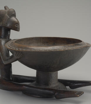 A wooden, bowl-bearing female figure is small compared to the vessel she sits and grips between her hands and long legs. She is nude with a shiny, naturally molded body and a face with simple, clear features. Her hair is represented by thin incisions.