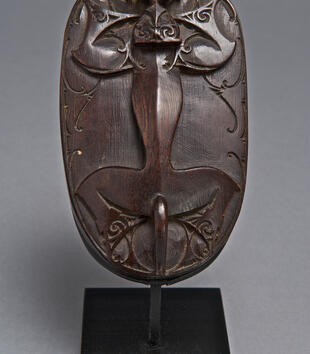 The top of a dark, wooden lid is carved with a depiction of an animal with a slender, serpentine waist and outstretched limbs. He has a small head with horns and blue, glass eyes. One of the beads is missing.