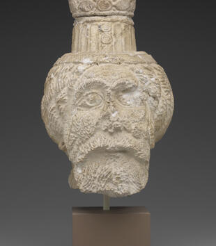 A limestone, life-sized head is carved with large open eyes, a beard, and bushy eyebrows. He wears a short and fluted cylindrical hat.
