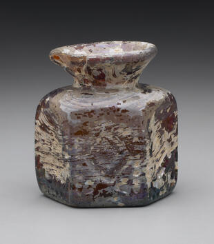A hexagonal flask made of blown glass is red and white in color. Its short neck supports a wide lip opening. A diamond is imprinted on one of the faces of the vase.