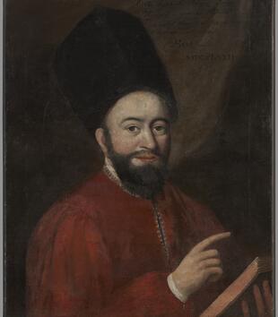 A painting represents Rabbi Raphael Haijm Isaac Karigal as a light-skinned man in red robes, a tall and boxy fur hat, and a dark beard. He holds a book in one hand and points to it with the other.