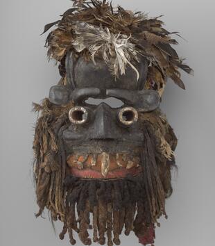 A dark wooden mask has geometric, white circular eyes, a pointed black nose, and jagged white teeth in an open mouth. Feathers are affixed to the top of the work and braided human hair hangs from the sides and chin of the face.