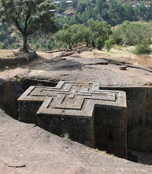 A photo looks down on a rock-hewn monolithic church of red volcanic tuff that has been carved down into a massive rock. The building has a cruciform shape and the top of the church is carved with a repeated pattern of nestled crosses.