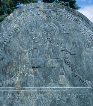 The top of a slate gravestone is carved with an image of a wide-eyed man preaching at a pulpit. A meandering line marks out an angel shaped pattern about the man that lends him a winged appearance.