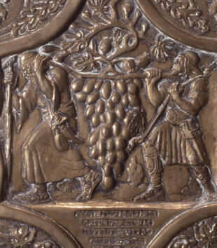 A metal relief depicts two men in robes with a pole over their shoulders, which they use to carry an oversized bushel of grapes.