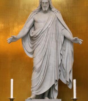 A white marble statue depicts a colossal figure of Christ with outstretched arms and downcast eyes. Long hair flows past his bearded face and down his shoulders. He is wrapped with a draping cloth that leaves his right arm and chest bare. 