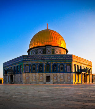 A photo captures the Dome of the Rock against a bright blue sky. The Islamic shrine consists of a golden dome atop a large octagonal base coated with blue and green mosaics.
