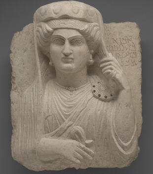 The bust of an elegant woman emerges in high relief from a limestone bust. She has large almond eyes with incised irises. She wears a tunic, a large broach on her shoulder, and a headdress. With one ringed hand, she holds back her veil.