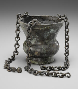 A bronze censer is vase-shaped, with a bulging mid-section and a wide lip. Three large chains are attached to the top of the censer for hanging the vessel.