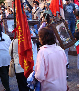 Tan-skinned women and children process down a street holding wooden frames on the back. The frames hold sculptures of saints.