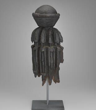 A wooden sculpture represents a highly abstract group of figures. The thin figures are carved in a row around a cylinder. The object is crowned with an orb-like ornament.