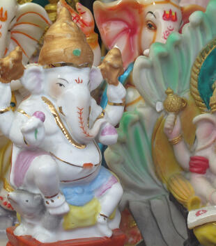 A collection of painted, plaster figurines depict the deity Ganesh in a variety of poses. The deity has the head of an elephant, four arms, and a golden crown. Ganeshs is shown sitting with a book, dancing, and sitting on a rat.