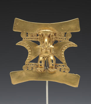 A gold pendant depicts a figure with the head of a bird and two large wings. He clutches a fish in his beak and his genitals are on view.