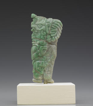 A green stone pendant depicting a standing man with a large headdress is rectangular shaped and richly carved. He has a large head, almond eyes, a craggy nose, and open mouth. His headdress has feathers and a beaded tassel. 
