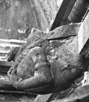 In a grayscale photo, a female stone torso with carved jewelry protrudes and swoops off a building. There is a jagged stone space where the figure's head should be.