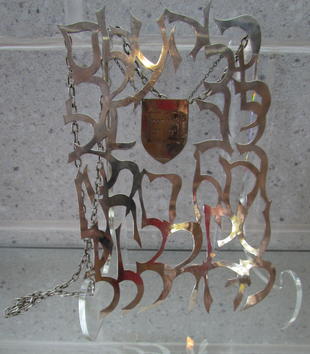 A silver metal sheet formed of swirling filigree frames a smaller metal shield. It hangs on a chair and is engraved with an image of an open book with Hebrew text. The whole piece hangs on more chains like a piece of jewelry.