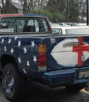 A pickup truck has been spray-painted like an American flag. The bed is blue with white stars and the front carriage has red and white stripes. The Jesus fish and a red cross are painted on the back. Religious bumper stickers are plastered over the trunk.