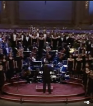 A still from a YouTube video features a choir, symphony orchestra, and conductor on the Carnegie Hall stage