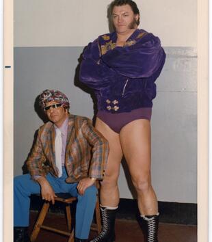 Film photograph of a wrestler, standing wearing a velvet jacket and underwear, and his manager, seated wearing a colourful suit and sparkly hat