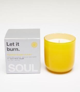 An unlit candle in a glossy, rounded, yellow glass container sits unboxed beside a silver boxed candle, which most prominently says "Let it burn." and "SOUL" in black, white, and yellow text. 