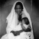 A black and white photo depicts a dark-skinned woman in a white veil cradling a swaddled baby close to her chest. She folds her hands in her lap and casts her eyes down. A nativity star is visible in the righthand corner of the photo.