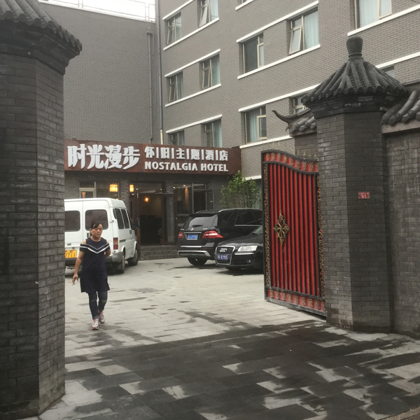 Red open gate off of a grey street, leading to a grey building with a brown awning that reads "Nostalgia Hotel" with a few cars parked in front. A woman walks out through the gate.