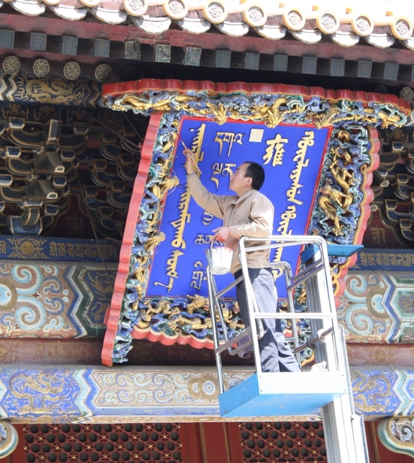 An artist with short hair stands on a scaffolding or crane structure and paints a blue, gold, and red sign with ornate carvings, set underneath an elaborate, gilded frieze. 