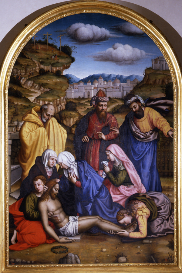 Light-skinned figures in bright drapery gather around a dead figure of Jesus laid on the ground. The group holds up the man's chest, and a woman kneels at his bleeding feet. A green landscape, white buildings, and blue sky form the painting's background.