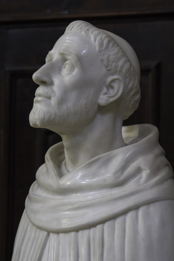 A male figure in shiny white marble gazes upwards with a placid expression. Viewed from the chest up, the man has a tonsure, slightly sunken cheeks, a scruffy beard, and friars robes.