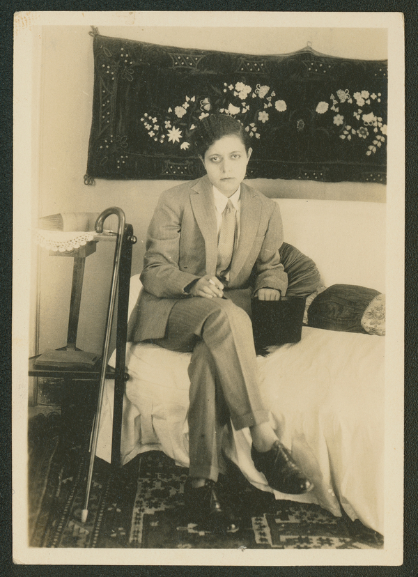In a sepia photo, a tan-skinned woman in a suit sits on a couch and stares out at the viewer. She holds a tall hat close while a cane rests to her side.