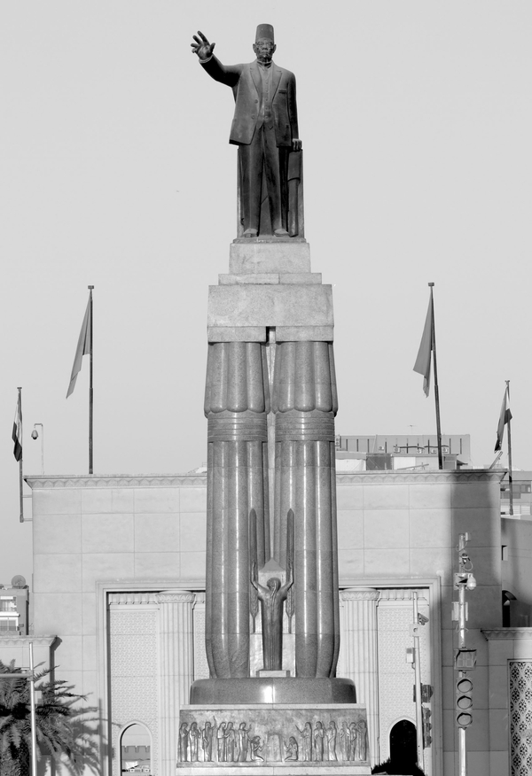A monumental bronze sculpture consists of a man orating atop a massive double column stand. The man raises his hand and wears a cap. Austere buildings and flags stand in the background.