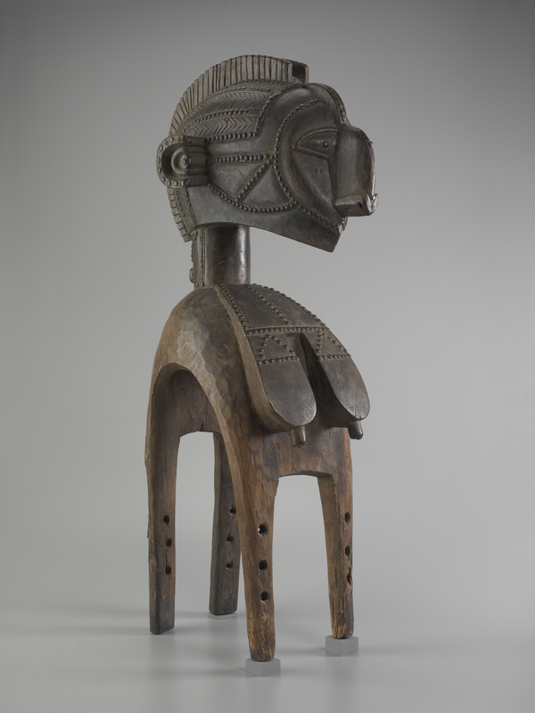 A large wooden figure has an oversized head with carved scarification. Flat breasts with carved, erect nipples and scarification flow over the sculpture's curved, four-legged base. The work's face has large ears and a rounded, protruding nose.