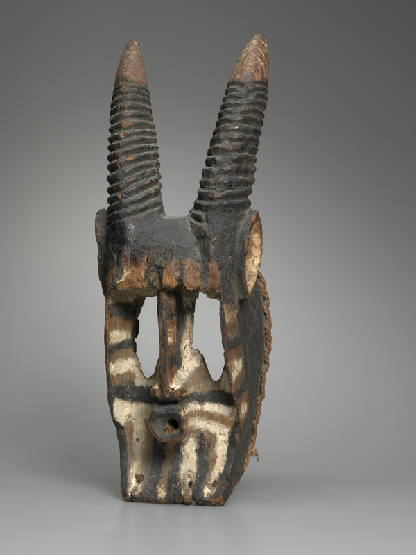 A carved wooden mask forms an elongated face with striped paint. It has long horns affixed on top, large eye holes, and and circular, puckered mouth.