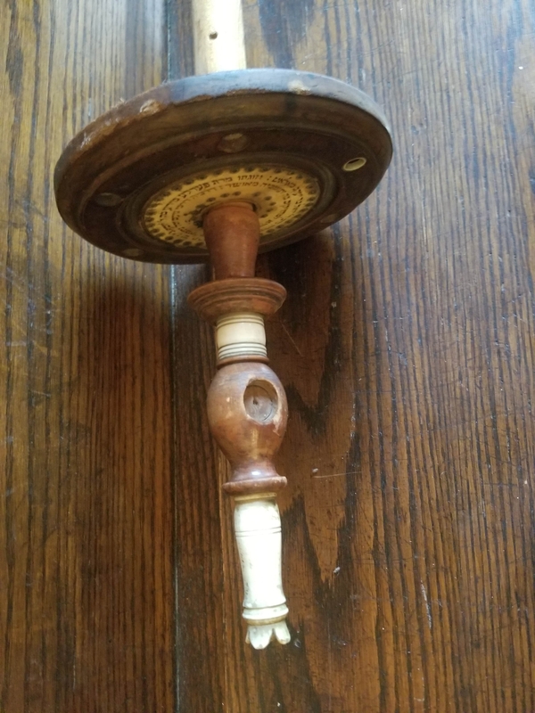 At the end of a bare, wooden Torah pole, there is a wooden disk with Hebrew lettering. It is topped by a wood and ivory ornament with empty niches for now-lost glass embellishments.