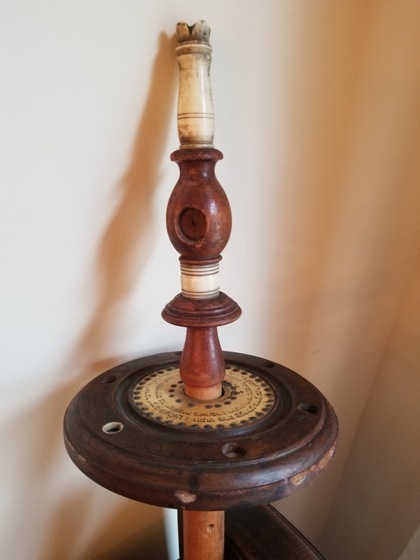 At the end of a wooden pole, there is a wooden disk decorated with Hebrew letters. A carved ivory and wood ornament is affixed on top. It has empty spaces for glass embellishments.