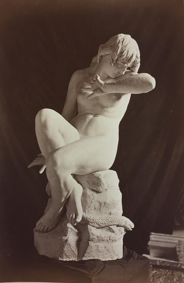 A clay model depicts a naturalistic nude of Eve cowering upon a rock. She moves to avert her face in the crook of her arm and crosses her legs to hide her body. A snake is visible curling around the rock near her feet.