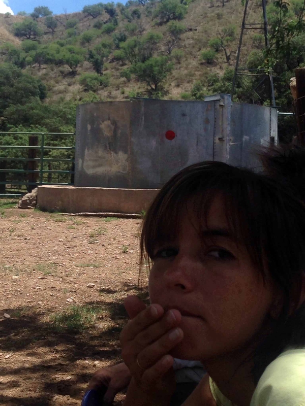 A tan-skinned young woman turns and faces the camera in the foreground of a photo of a silver, outdoor water tank. The tank is spray-painted at the center with a red dot.
