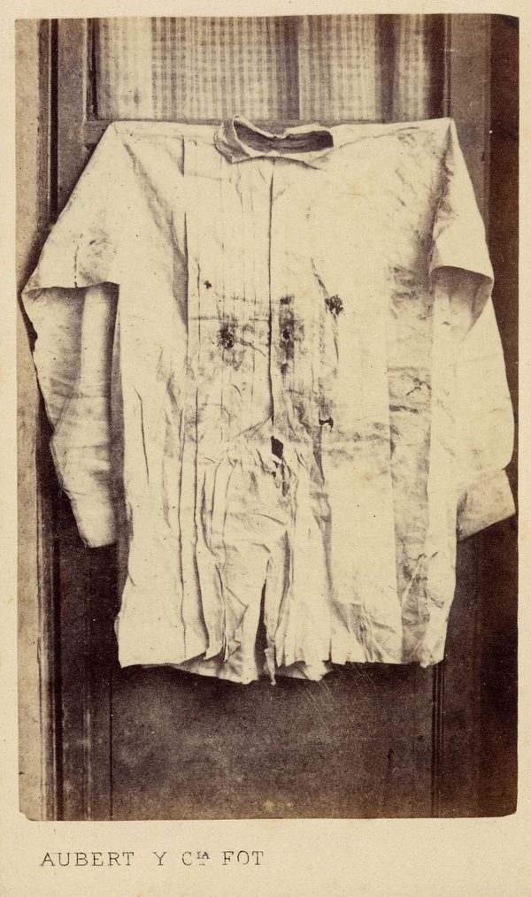 This sepia photo records a bloodied shirt pieced with bulletholes. The shirt is pinned to the crossbeam of a window and centered in the photo.