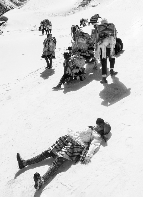 A black and white photo shows Peruvian pilgrims of all ages and genders travelling through the snow on a mountainside. A young girl laughs and lays down in the whiteness. She wears a striped skirt and lacy top.