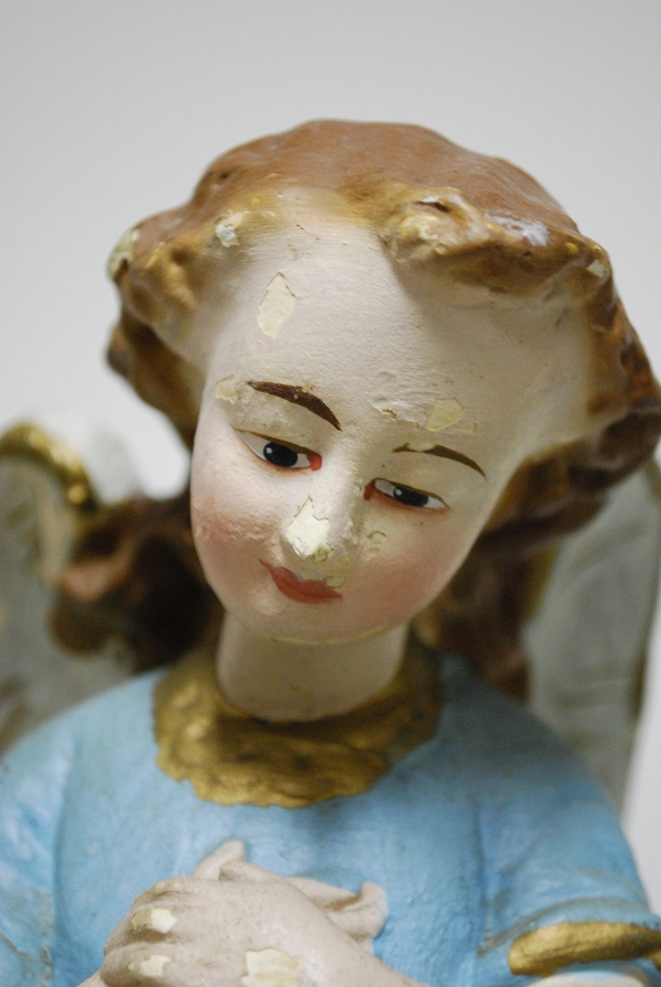 A close-up photo of the face of a light-skinned angel with delicate red lips, thin brown eyebrows, and downcast eyes has paint chipping from the surface.