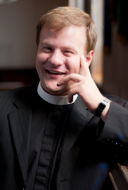 A middle-aged, light-skinned man wearing a clerical collar smiles and rests his head on his hand.