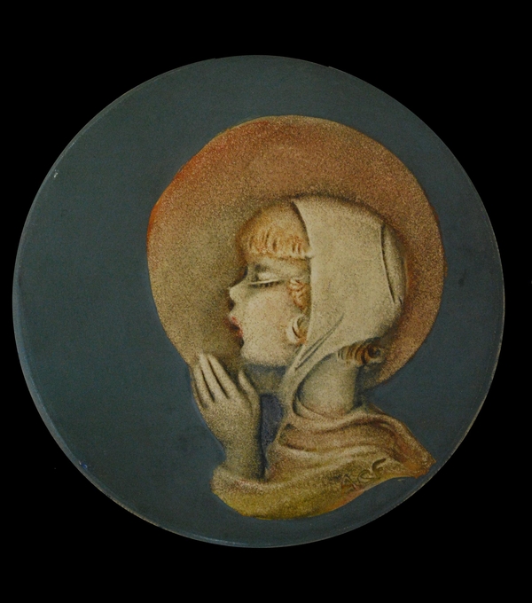 A round blue plastic disc is painted with a profile portrait of a young, light-skinned girl. A disc of light encircles her peaceful face as she clasps her hands together in prayer. She wears a white cap over her blonde hair.  