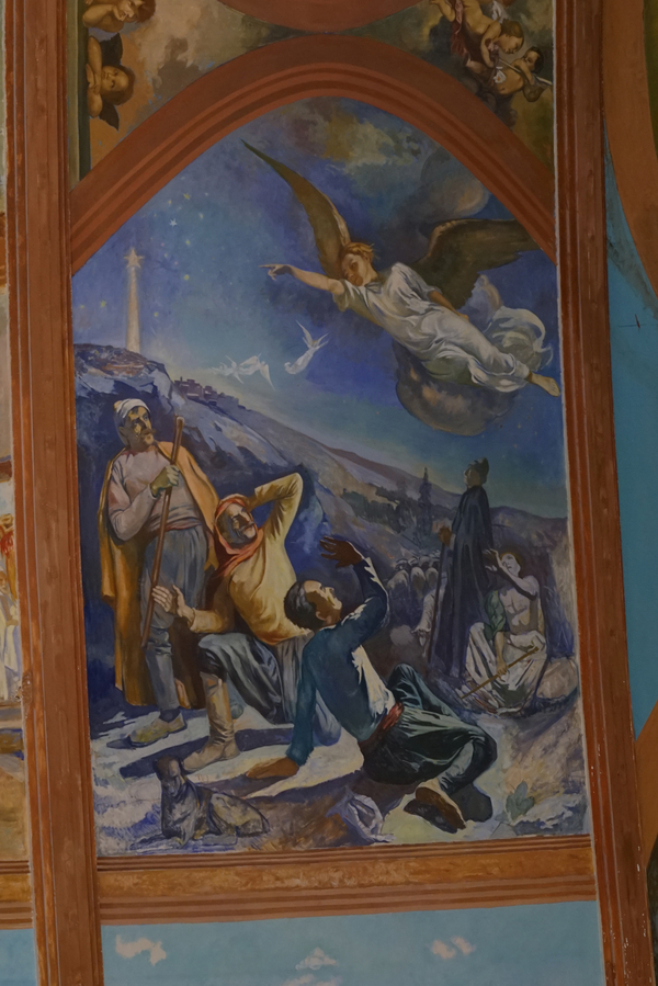 A section of a church mural painting depicts an angel flying in a dark blue sky above startled shepherds. A shining star hovers above a mountain in the background.