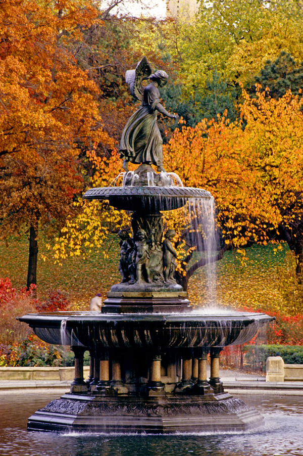 A two-tiered water fountain stands against a backdrop of autumn trees. A bronze angel tops the second tier. She wears a flowing dress and steps forward with her left foot. Chubby putti decorate the base of the second tier.