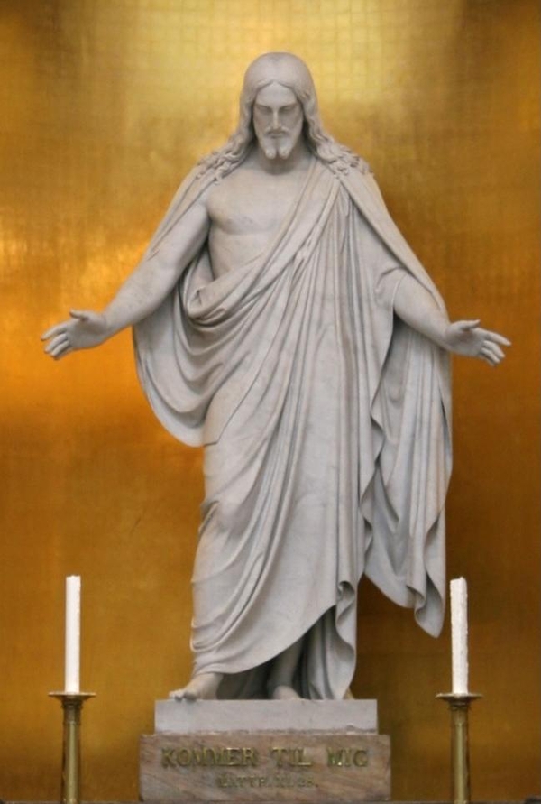 A white marble statue depicts a colossal young male figure with outstretched arms and downcast eyes. Long hair flows past his bearded face and down his shoulders. He is wrapped with a cloth that leaves his right arm and chest bare. 