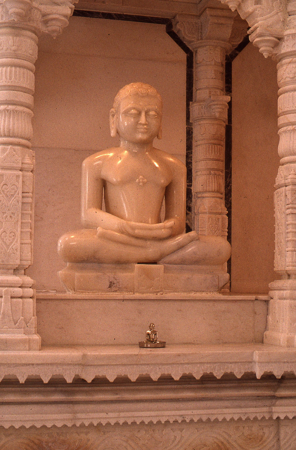 A shiny, light-colored stone statue of a large figure with long earlobes, serene eyes, and a bare chest sits crosslegged and with arms folded. The figure is in an ornately carved shrine of the same stone. 